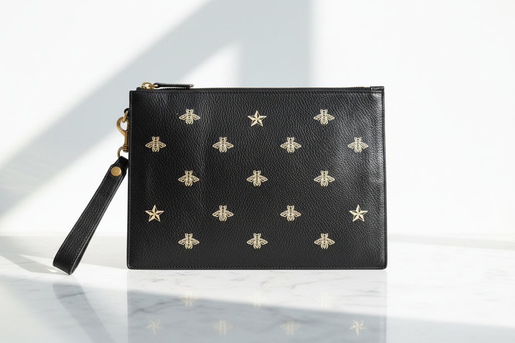 Elegant bright editorial view of Gucci black leather wristlet clutch with gold embossed bee and star motifs on white marble